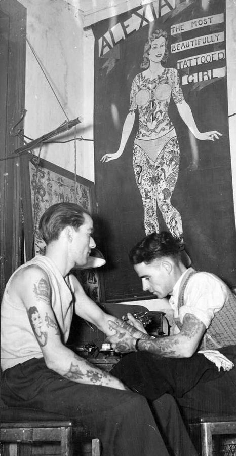Dois tatuadores em um estúdio, com um cartaz de mulher tatuada ao fundo